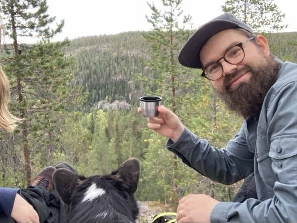 I man is raising a thermos cup of coffee into the camera while sitting in a hilly forested nature setting, next to a dog and a woman.