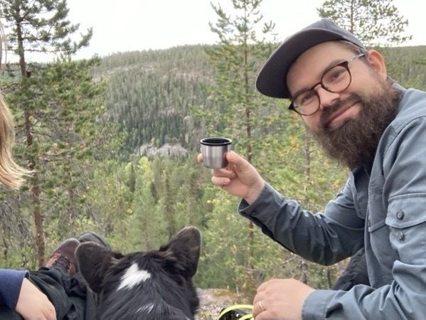 IMG_5107.jpeg I man is raising a thermos cup of coffee into the camera while sitting in a hilly forested nature setting, next to a dog and a woman.
