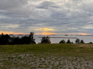 A photo of a cloudy sky over the coast near Oulu. In the middle of the photo, the sun is breaking through the otherwise grey-whitish clouds turning the sky orange-yellow. A ray of light seems to focus on one of the little islands scattered on the horizon.